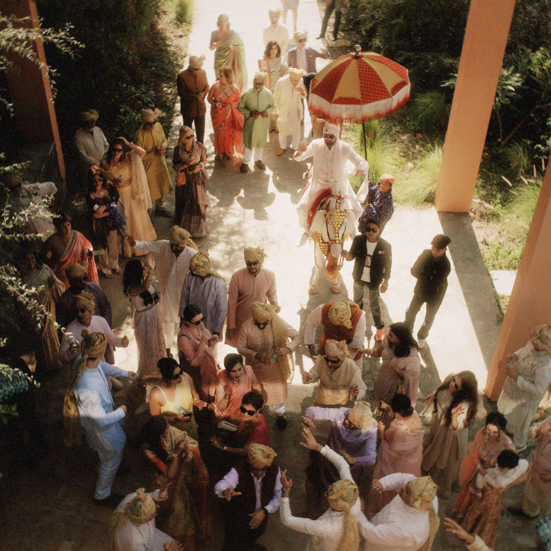 Wedding guests in colourful outfits gathered under parasols at a Baraat